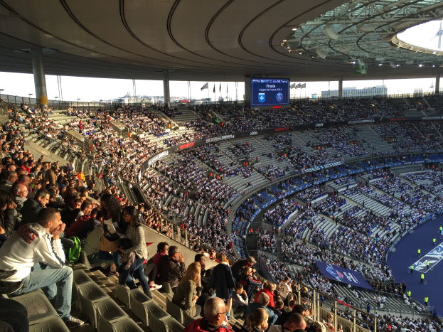 Eine große Menschenmenge sitzt im Allianz Arena in München, Deutschland, und schaut ein Fußballspiel, mit einer Bühne auf der rechten Seite, Fahnen, Stangen und einem Bildschirm im Hintergrund und dem Himmel oben sichtbar.