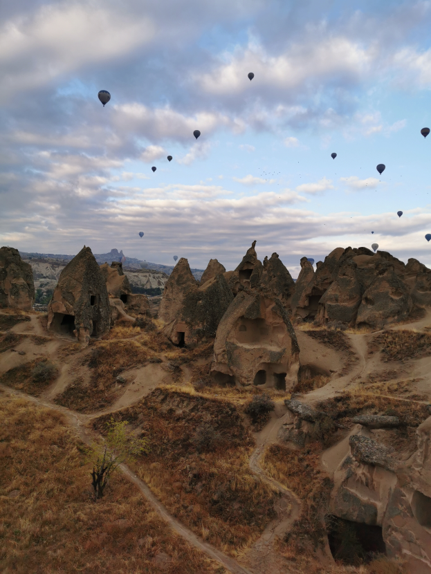 Heißluftballons fliegen über steinige Hügel in Kappadokien, Türkei, mit Fallschirmen und Wolken im Hintergrund.