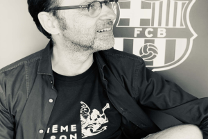 Ein Mann in einer Brille und einem schwarzen Hemd lächelt vor einem schwarz-weißen FC Barcelona-Logo an einer Wand.