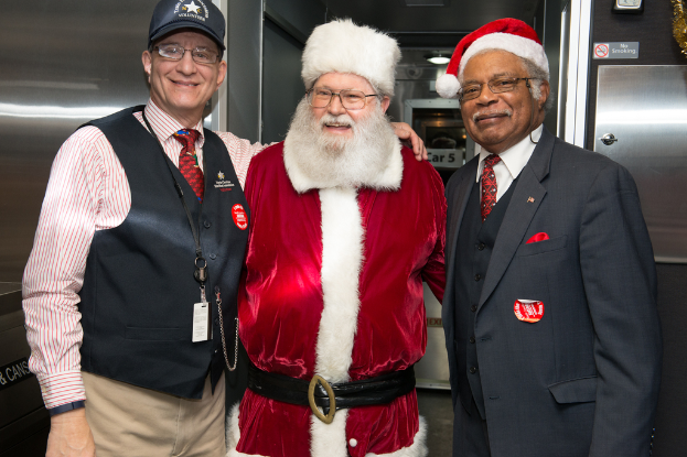 Eine Gruppe von Menschen mit Brillen, Mützen, Krawatten, ID-Karten und Abzeichen, darunter eine Person in einem Weihnachtskostüm, die vor einer Stahlwand mit Lichtern und Schildern steht.