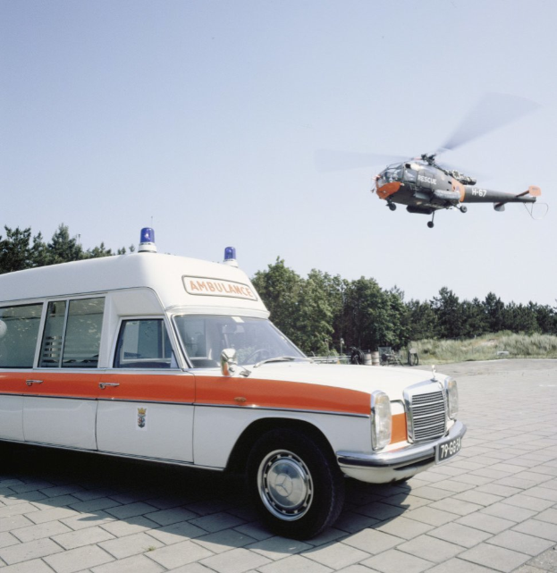 Ein Rettungswagen steht vor einem Hubschrauber, mit Bäumen im Hintergrund und einem klaren blauen Himmel darüber.