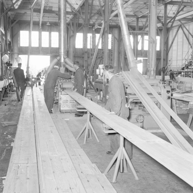 Schwarz-weiß-Foto von Männern, die an einer Maschine in einem vollgestellten Fabrikgebäude arbeiten, das mit Holzplanken, Pfählen, Lampen und Fenstern gefüllt ist.
