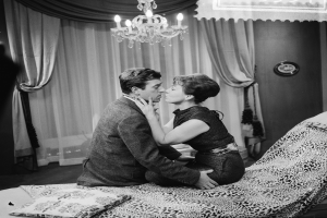 Ein Mann und eine Frau umarmen und küssen sich zärtlich auf einem Bett mit Kissen und einer Decke, unter einem Kronleuchter in einem sanft beleuchteten Raum mit Vorhängen.