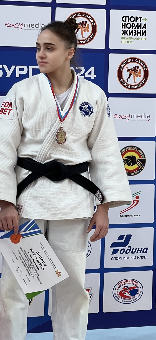 Frau in weißem Judo-Anzug mit schwarzem Gürtel auf einem Podest, die eine Medaille und ein Zertifikat hält, mit einem Logo-Schild im Hintergrund.