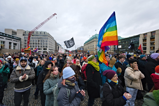 Große Gruppe von Menschen bei einer LGBTQ+-Rechtsdemo in Berlin mit Fahnen und Spruchbändern vor einem Gebäude, mit Kränen und bewölktem Himmel im Hintergrund.