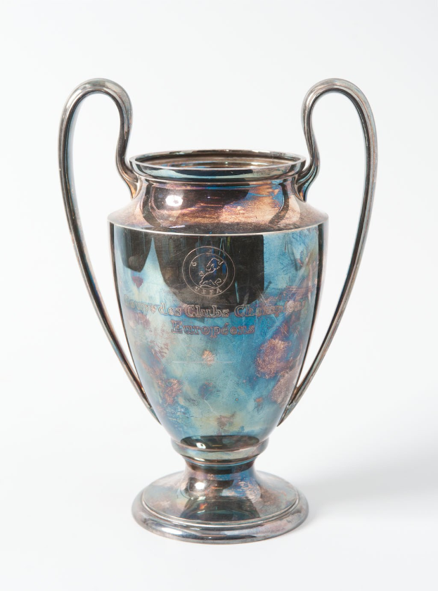 Der UEFA-Champions-League-Pokal, ein stahlerner Becher mit zwei Handles, blau und silbern, mit der Aufschrift "UEFA Champions League" auf einem weißen Hintergrund.