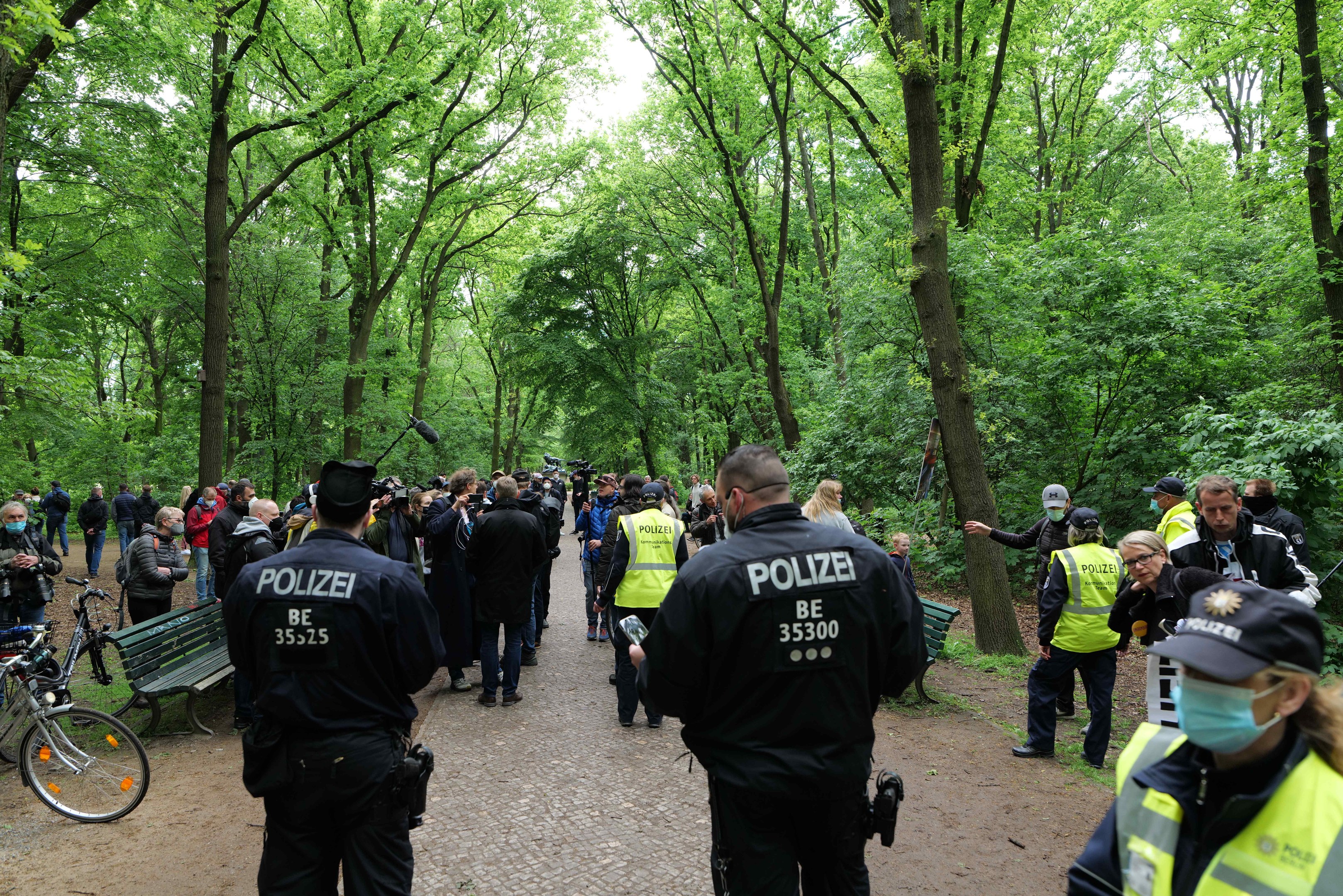 Eine Gruppe von Polizeibeamten steht vor einer Menschenmenge, mit Fahrrädern und einer Bank im Vordergrund, Bäumen und Himmel im Hintergrund, während einer Anti-Terror-Demonstration in Berlin.