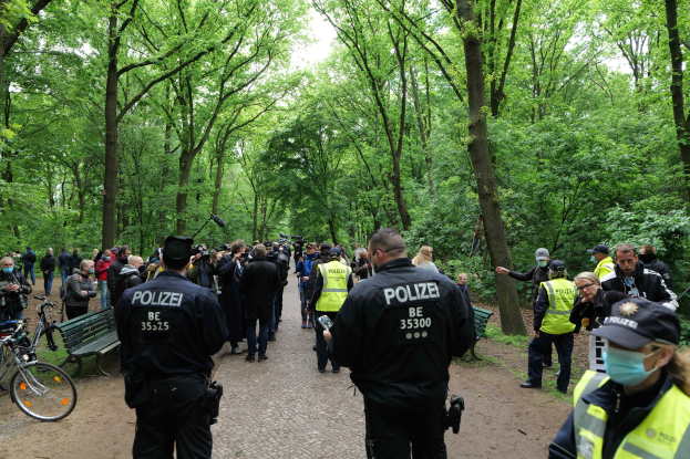 Eine Gruppe von Polizeibeamten steht vor einer Menschenmenge, mit Fahrrädern und einer Bank im Vordergrund, Bäumen und Himmel im Hintergrund, während einer Anti-Terror-Demonstration in Berlin.