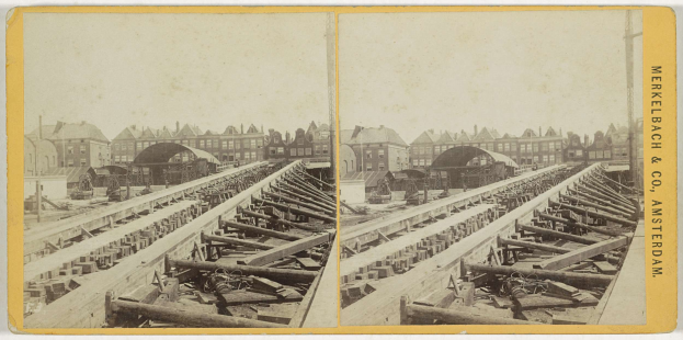 Altes Schwarz-Weiß-Foto einer Zugstrecke mit Gebäuden im Hintergrund, Text 'Merkelbach & Co, Co. Amsterdam' auf der rechten Seite und verstreute hölzerne Gegenstände im Vordergrund.
