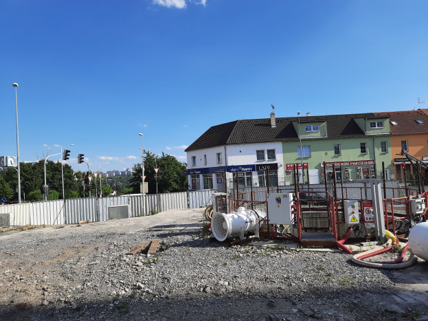 Eine Baustelle mit verstreuten Rohren und Geräten, umgeben von Gebäuden, Laternen, Ampeln, einem Zaun, Bäumen und einem bewölkten Himmel.