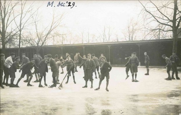 Ein Schwarz-Weiß-Bild einer Gruppe von Männern, die Hockey auf einem Eisplatz spielen, mit Bäumen und einer Hütte im Hintergrund und Text oben.