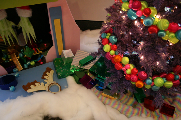 Eine Indoor-Szene mit einer Wand im Hintergrund, einem Baumwoll- und Wolltuch am Boden, einem geschmückten Weihnachtsbaum rechts, Geschenkboxen und Dekorationsgegenständen in der Mitte und einer Hütte mit Dekorationen und Baumwolle oben links.