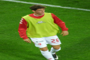 Ein Mann in einem gelben und roten T-Shirt läuft auf einem Fußballfeld mit weißen Begrenzungslinien, trägt Fußballschuhe.