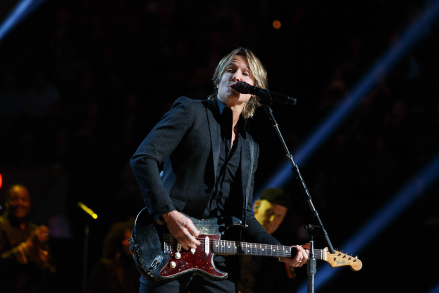Keith Urban performs on stage at the Joint in Las Vegas, playing a guitar with a microphone in front of him and musicians in the background, under dark lighting.