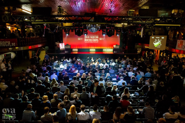Eine große Menschenmenge sitzt auf Stühlen vor einer Bühne mit Bildschirmen und Bannern, die von Scheinwerfern beleuchtet wird und von Geländern umgeben ist, mit einem Wasserzeichen in der linken unteren Ecke, in Erwartung des Beginns eines Online-Marketing-Rockstars-Events.