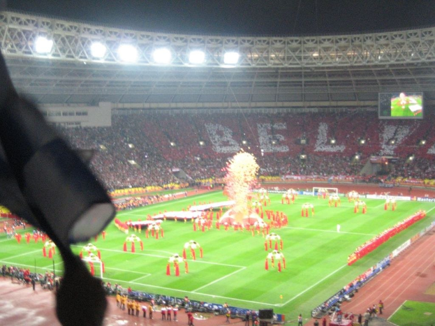 Ein Stadion mit Flutlicht, Eisengittern, Zuschauern, stehenden Personen auf dem Boden, Werbetafeln, Anzeigetafeln, eine Überwachungskamera, Fahnen und Dekorationen.