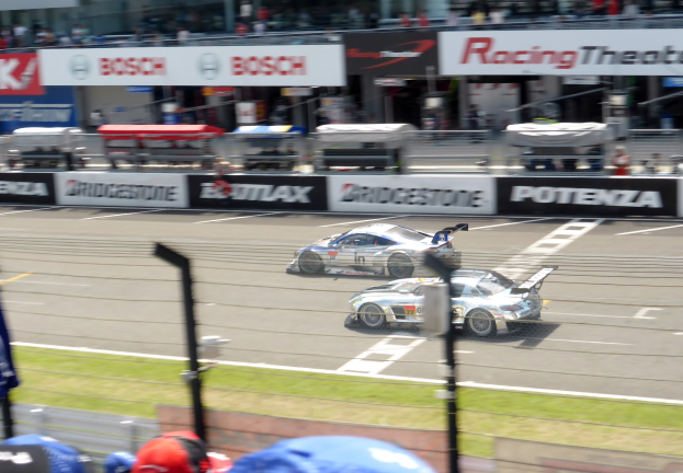 Zwei Rennautos auf einer Rennstrecke mit Zuschauern im Hintergrund, einschließlich Menschen in Mützen und Werbetafeln.