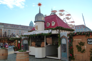 Ein kleines Gebäude mit einem roten Dach und einem Riesenrad davor, umgeben von Menschen und Festdekorationen bei einem Oktoberfest in München, Deutschland.