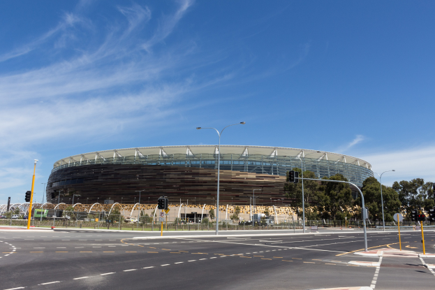 Großes Stadion mit starkem Verkehr davor, umgeben von Straßeninfrastruktur einschließlich Pfosten, Laternen, Signalen, Schildern, Geländern, Bäumen und einer bewölkten Himmel.