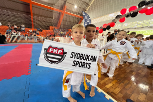 Eine Gruppe von Kindern steht auf einer blauen und roten Matte und hält ein Banner mit der Aufschrift "Federação de Karatê Paulista". Im Hintergrund sind Menschen, ein Netz, Luftballons, Lichter und eine Wand zu sehen.