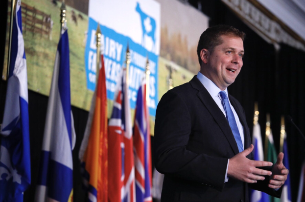 Ein Mann im Anzug spricht an einem Podium mit Mikrofonen, umgeben von Fahnen und einem Bildschirm, wahrscheinlich bei der Ankündigung einer Regierungszahlung von 1,5 Milliarden Dollar für die Milchindustrie.