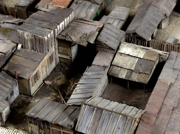 Ein Modell eines kleinen Dorfes mit verstreuten Metallhütten, Erde als Boden und einigen Bäumen im Hintergrund.