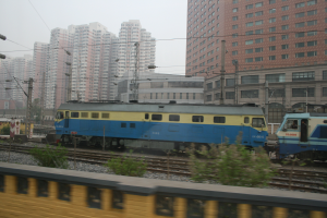 Zwei Züge fahren auf Schienen neben hohen Gebäuden, mit Strommasten, Drähten, Pflanzen, Steinen und einem Zaun im Vordergrund und dem Himmel im Hintergrund.