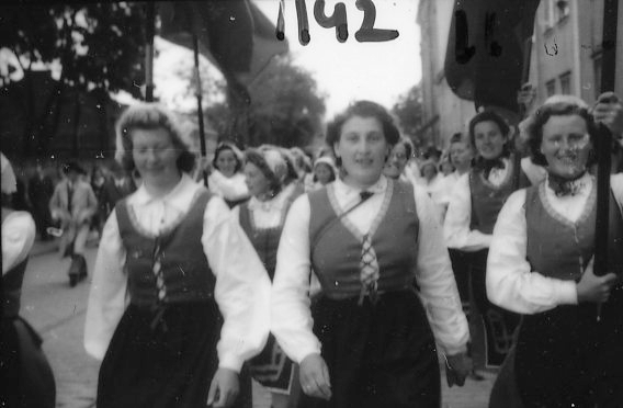 Eine Gruppe von Frauen in traditioneller bayrischer Tracht spaziert strahlend durch eine Straße, umgeben von Bäumen und Gebäuden, auf einem Schwarz-Weiß-Bild.