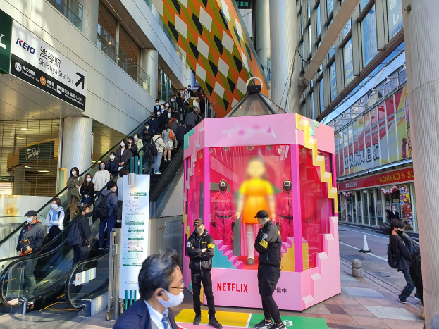Eine Gruppe von Menschen steht vor einem Gebäude mit Glaswänden, Säulen und Texttafeln, einige tragen Masken, während eine Rolltreppe mit Menschen zu sehen ist. In der Mitte steht ein pinker Kasten mit Schaufensterpuppen und der Aufschrift "Netflix's new Netflix booth in Tokyo."