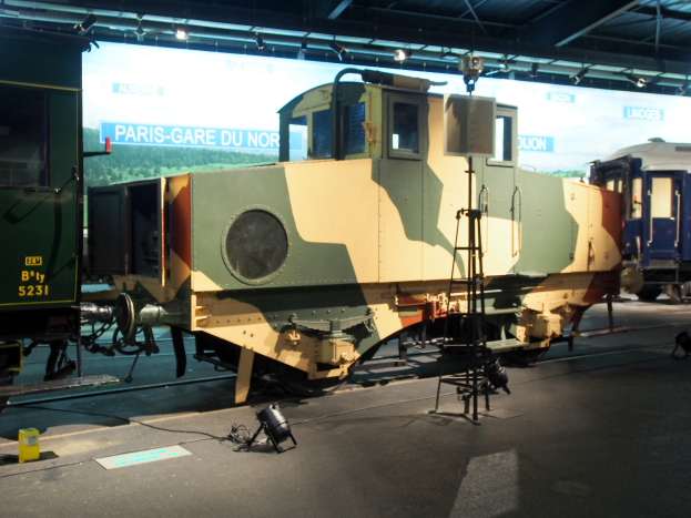 Eine Lokomotive in einem Museum ausgestellt, umgeben von anderen Zügen, mit einer Leiter, Lampen und verschiedenen Gegenständen, die auf dem Gleis verstreut sind, und einem Banner mit Text im Hintergrund, unter einer beleuchteten Museumsdecke.