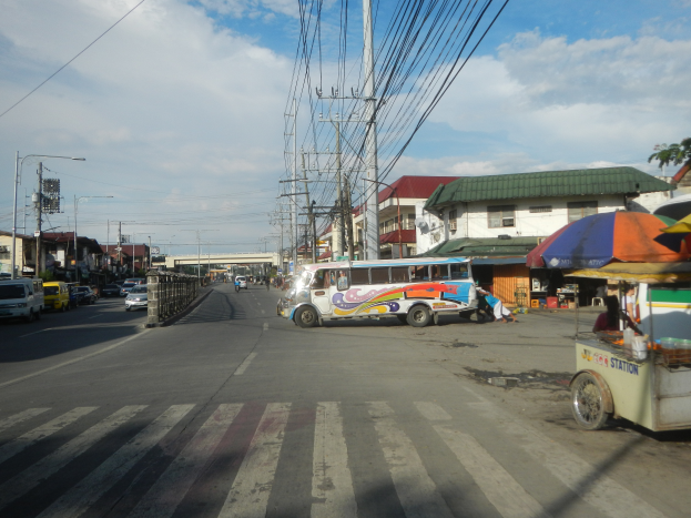 Eine Stadtstraße mit parkenden Fahrzeugen, darunter ein Bus, Strommasten mit Drähten, Laternen, Gebäude mit Fenstern, eine Brücke und ein bewölkter Himmel im Hintergrund.