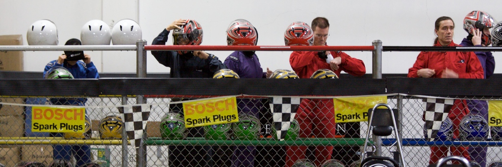 Ein Maschendrahtzaun mit Plakaten und Fahnen, mit mehreren Personen im Hintergrund, einige tragen Helme, und zusätzliche Helme auf einem Ständer, alles vor einer Wand.