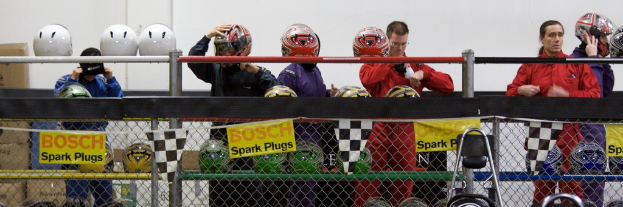 Ein Maschendrahtzaun mit Plakaten und Fahnen, mit mehreren Personen im Hintergrund, einige tragen Helme, und zusätzliche Helme auf einem Ständer, alles vor einer Wand.