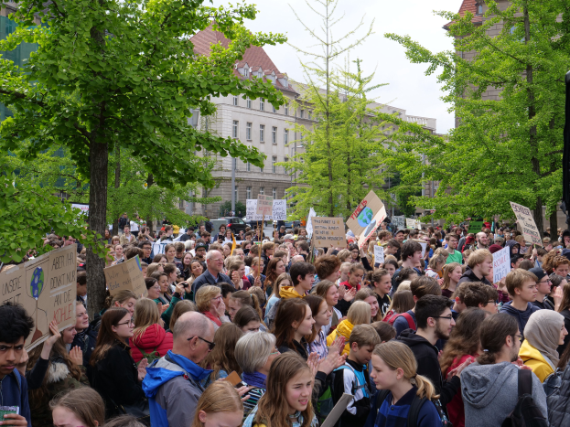 Eine große Menschenmenge mit Schildern steht vor einem Gebäude in Berlin, mit Bäumen, Fahrzeugen und einem Lautsprecher, unter einem klaren Himmel.