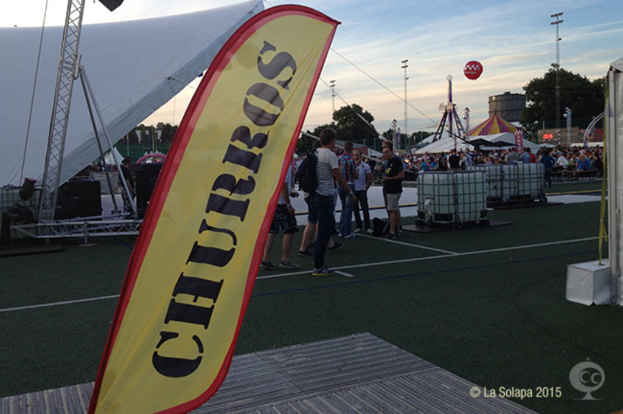 Ein buntes Volksfest-Szenario mit Zelten, einem zentralen Mast, Menschen, Bäumen, Strommasten, Straßenbahnen auf der Straße und einem Eisengestell auf der linken Seite unter einem klaren Himmel.