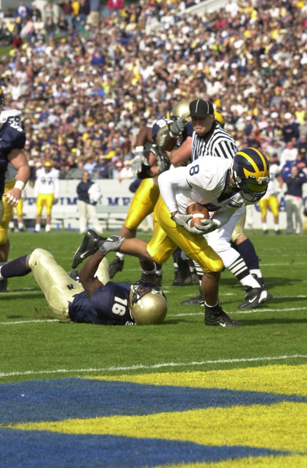 Ein Fußballspieler in Uniform rennt mit dem Ball, während er von Zuschauern in Sportbekleidung und Helmen umgeben ist, wobei Stadiontafeln, Geländer und andere Elemente im Hintergrund sichtbar sind.