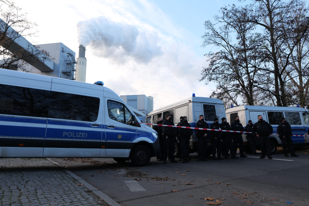 Polizeibeamte vor einem weißen und blauen Van auf einer Straße mit Bäumen, Gebäuden und einer Fabrik im Hintergrund unter einem blauen und weißen Himmel.