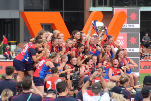 Eine Gruppe von Frauenfußballerinnen in blauen und roten Uniformen feiert mit einem Pokal vor einem jubelnden Publikum auf einem Rasenfeld, mit Zuschauern und beschrifteten Tafeln im Hintergrund.