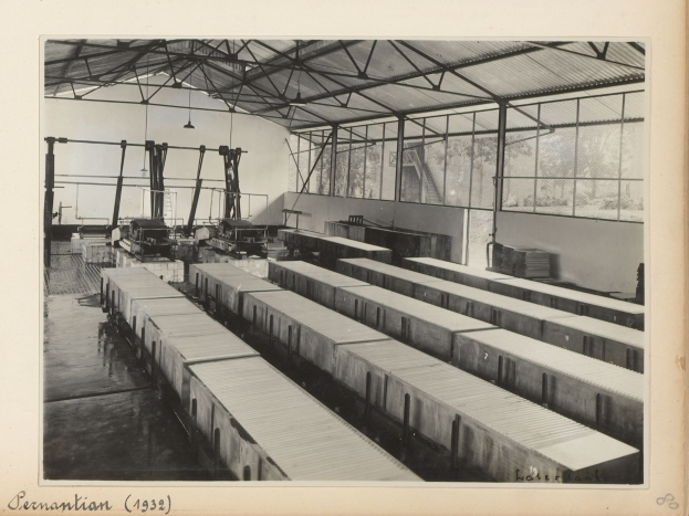 Schwarzes und weißes Foto einer Industrieanlage mit einem Zug auf Schienen, umgeben von Maschinen, Polen, Lampen und Glasfenstern, mit Text unten.