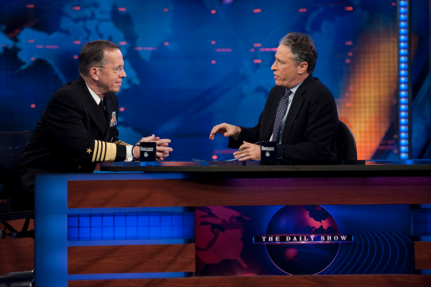 Zwei Männer in schwarzen Blazern, Hemden und Krawatten sitzen an einem Tisch mit Mikrofonen dazwischen und führen ein Gespräch vor einem Fernseher und einem blauen Hintergrund.