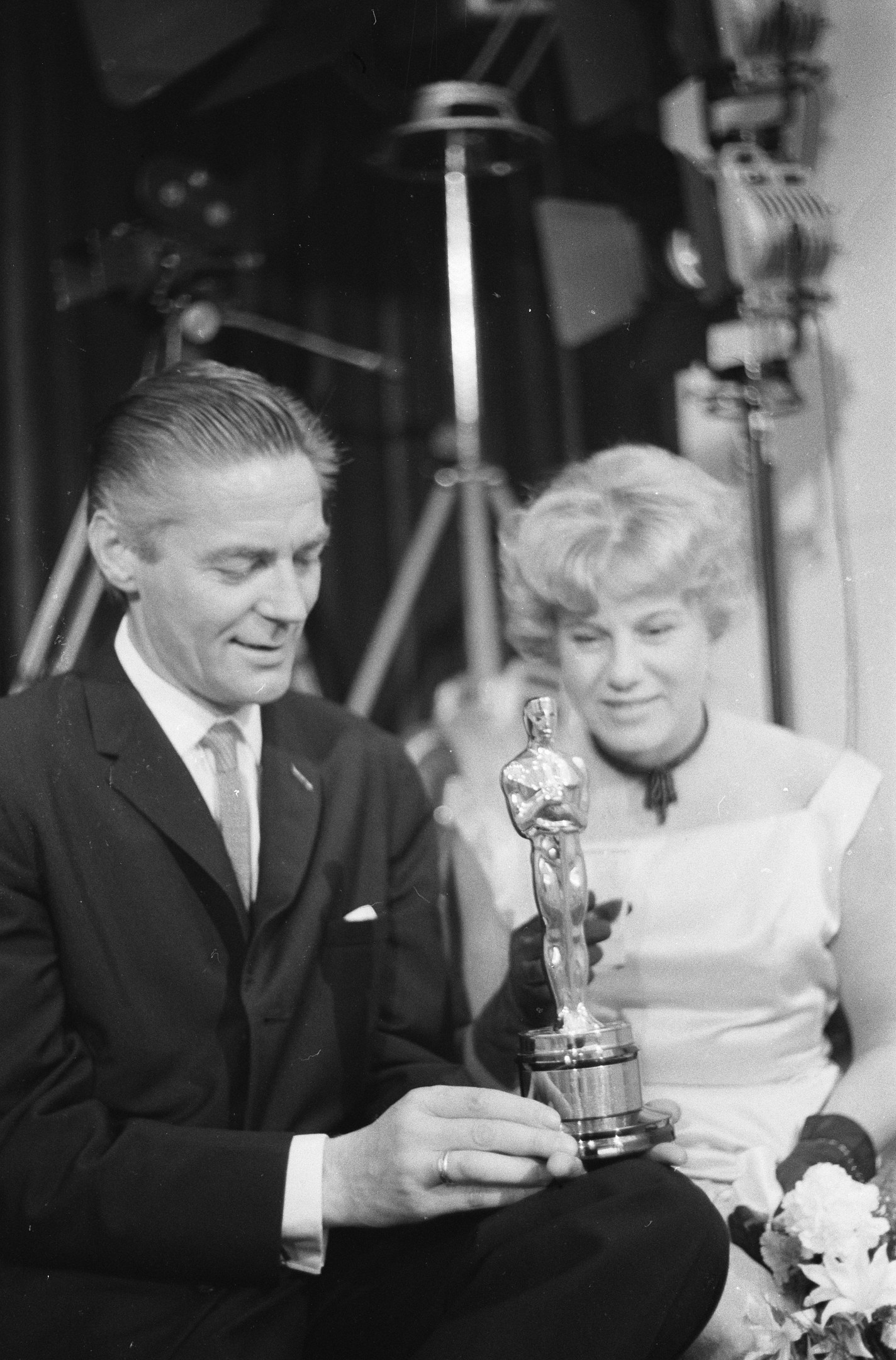 Ein Schwarz-Wei├č-Bild eines formell gekleideten Mannes, der eine Oscar-Statue hölt und eine Frau, die einen Blumenstrau├č hölt, nebeneinander sitzend mit beleuchtetem Hintergrund.