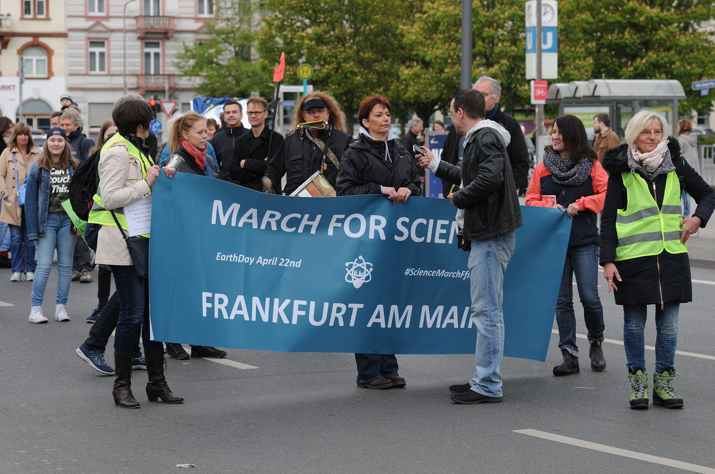 Gruppe von Menschen marschiert die Straße entlang und hält ein "March for Science Frankfurt am Main"-Schild mit Bäumen, Pfählen, Schildern und Gebäuden im Hintergrund unter einem klaren blauen Himmel.