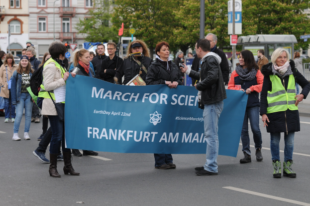 Gruppe von Menschen marschiert die Straße entlang und hält ein "March for Science Frankfurt am Main"-Schild mit Bäumen, Pfählen, Schildern und Gebäuden im Hintergrund unter einem klaren blauen Himmel.