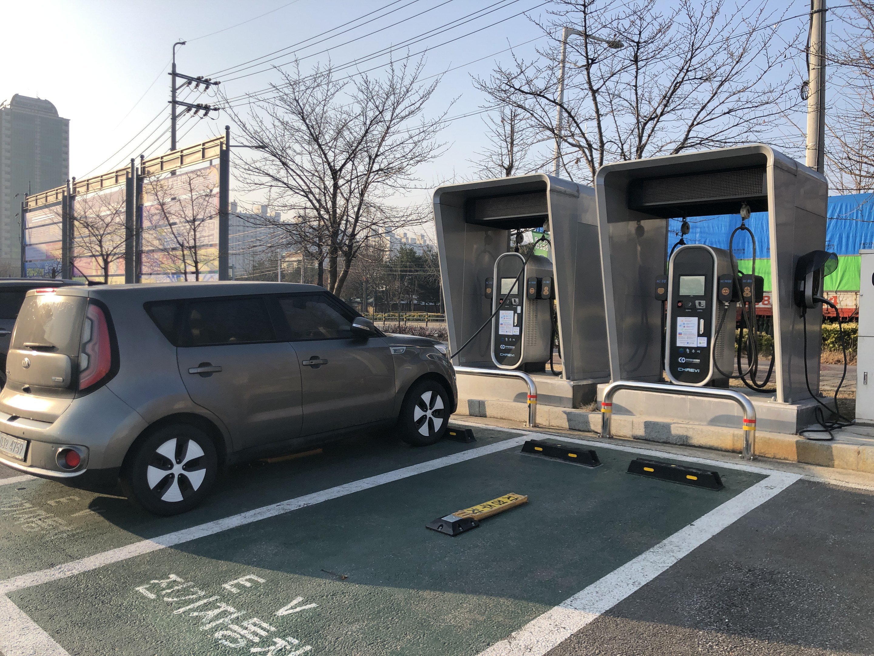 Elektrisches Auto-Ladestation auf einem Parkplatz mit Bäumen, Gebäuden, Strommasten und einem klaren Himmel.