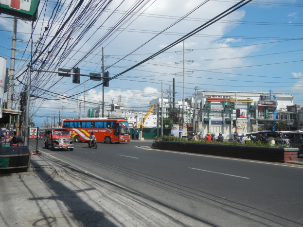 Eine belebte Stadtstraße mit Fahrzeugen, Ampeln, Strommasten, Gebäuden, Bäumen und einem bewölkten Himmel.