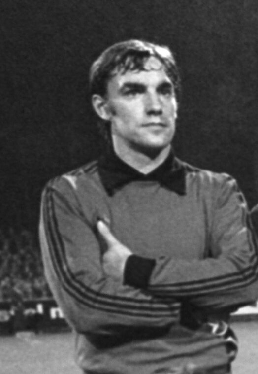 Schwarzes und weißes Foto eines Mannes auf einem Fußballfeld mit verschränkten Armen, umgeben von einer Gruppe von Menschen im Hintergrund.