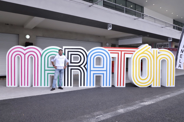 Ein Mann steht am Start eines Marathons vor einem großen "Marathon"-Schild, mit einem Banner rechts daneben und einem Gebäude mit Lichtern, Geländern und Fensterläden im Hintergrund.