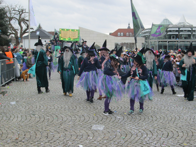Gruppe von Menschen in Hexenkostümen, die eine Kopfsteinpflasterstraße entlanggehen, schwarze und lila Kleider und Hüte tragend, einige Fahnen haltend, mit einem Geländer auf der linken Seite und Gebäuden, Bäumen und einem klaren blauen Himmel im Hintergrund.