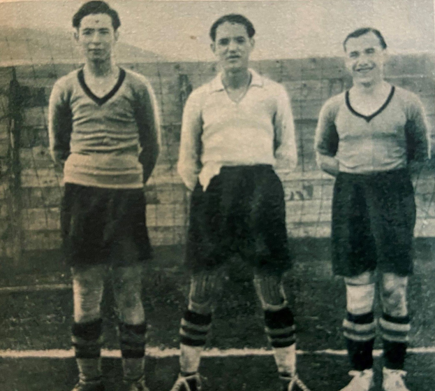 Drei Männer in Fußballtrikots und Fußballschuhen stehen nebeneinander auf einem Fußballfeld mit einer Wand im Hintergrund.