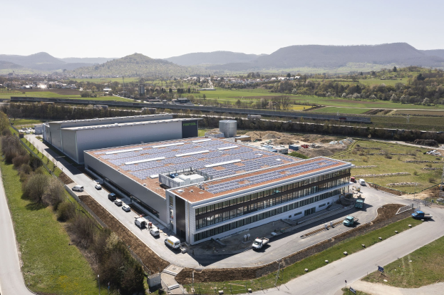 Luftaufnahme eines großen Gebäudes mit Solarpanelen auf dem Dach, umgeben von Fahrzeugen, Bäumen, Gras und Hügeln unter einem klaren blauen Himmel, identifiziert als Deutschlands erste solarbetriebene Fabrik.